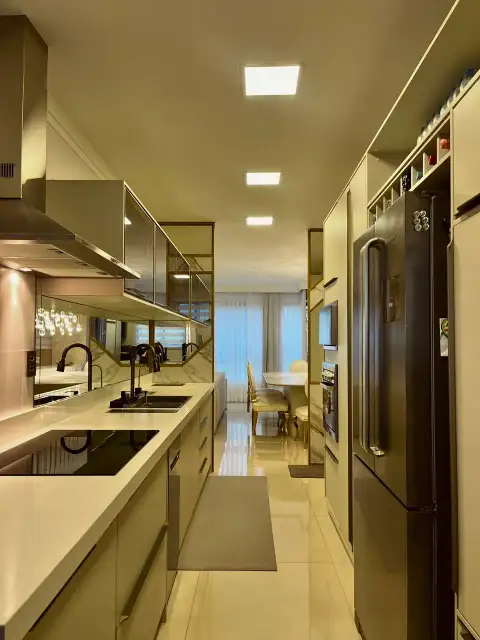 Cozinha em corredor com acabamento dourado e vista para sala de jantar iluminada