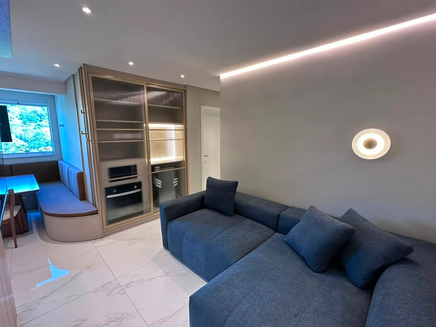 Sala de estar com sofá em tecido azul-marinho, luz de canto dourada e parede lisa para painel